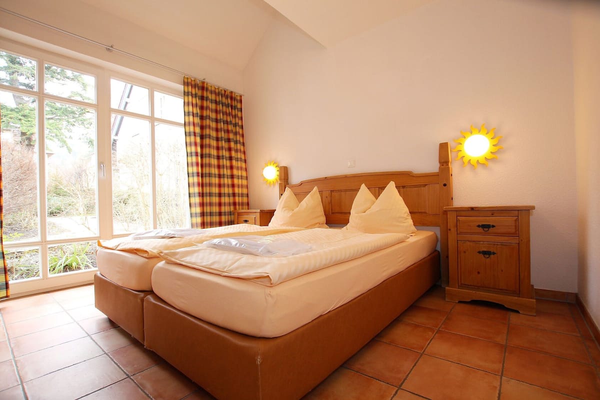A bedroom with a bed, a nightstand, a wall-mounted lamp, and a window with curtains.
