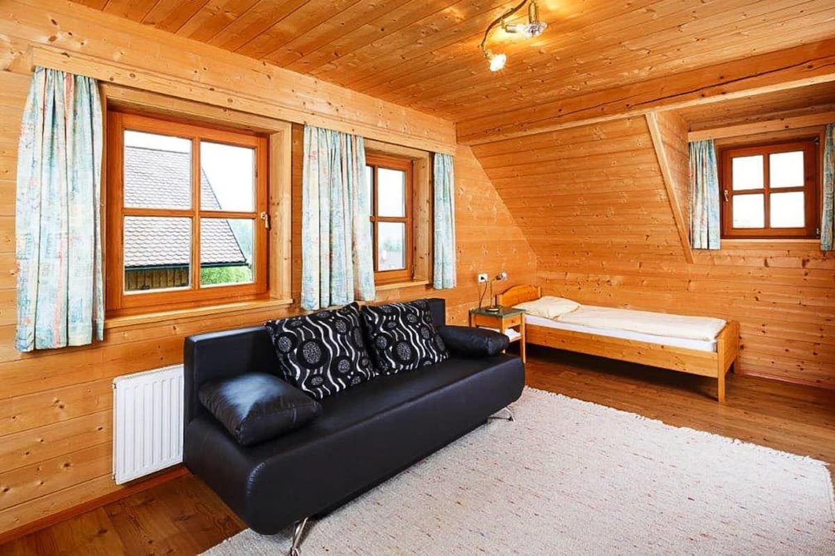 A cozy wooden cabin with a black leather sofa, a small wooden bed, and two windows with curtains.