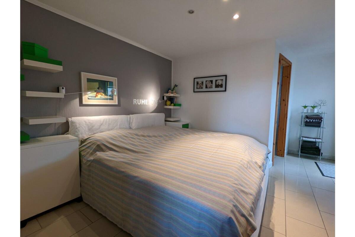 Ein Schlafzimmer mit Bett, Nachttischen, einer Kommode, einem an der Wand befestigten Regal mit Dekoration und einer Tür zu einem anderen Raum.