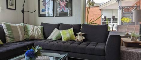 A living room with a dark sofa, a glass coffee table, and a teddy bear. There are framed pictures on the wall and a window with a view of a brick building.