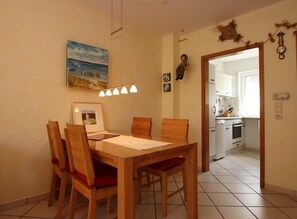 A dining area with a wooden table and chairs, a painting on the wall, and a kitchen visible through an open door. - Reihenhaus, Dornumersiel (Dornum)