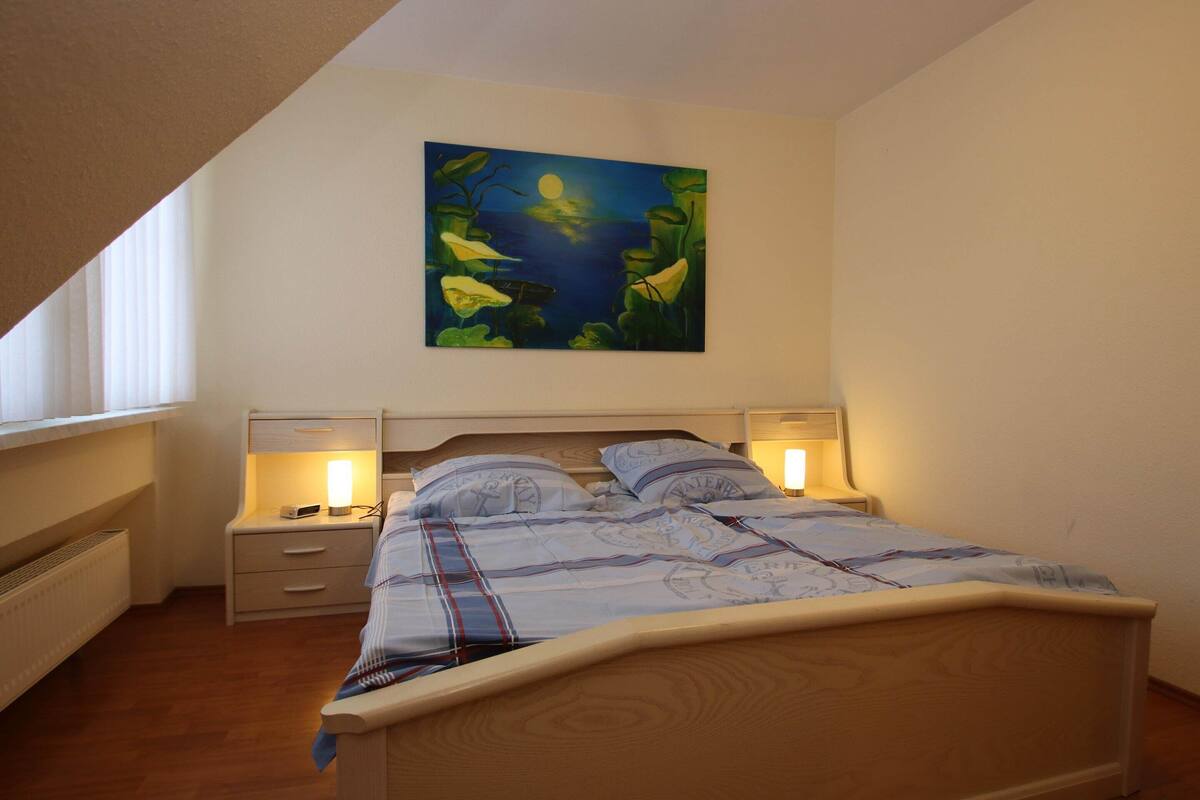 A bedroom with a bed, bedside tables, a nightstand, a painting on the wall, and a window.
