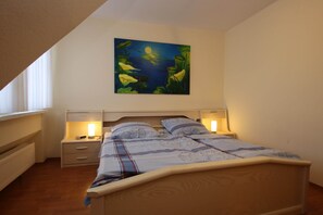 A bedroom with a bed, bedside tables, a nightstand, a painting on the wall, and a window. - Reihenhaus, Dornumersiel (Dornum)