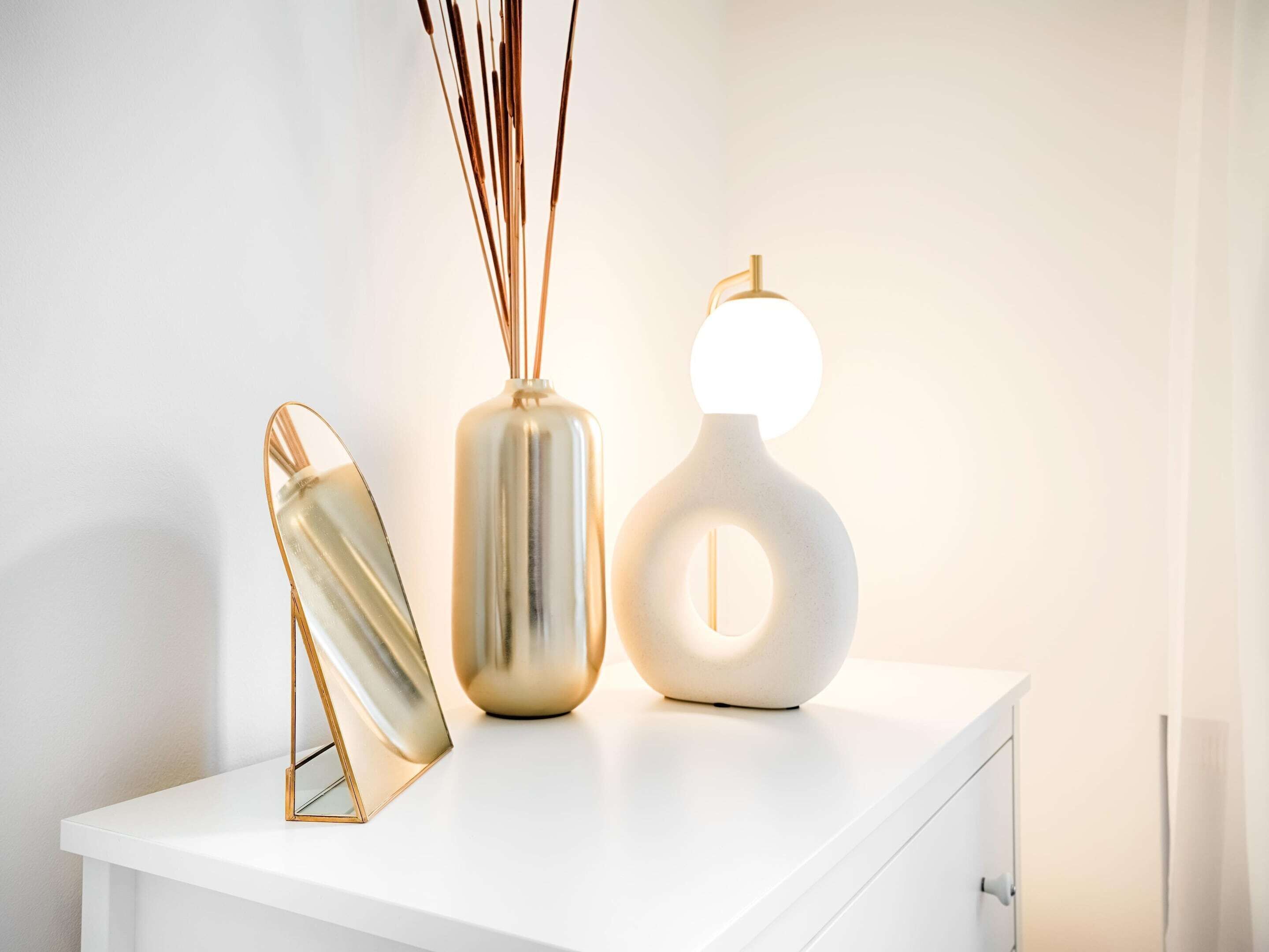A white bedside table with a mirror, a cylindrical vase with reeds, and a modern lamp.
