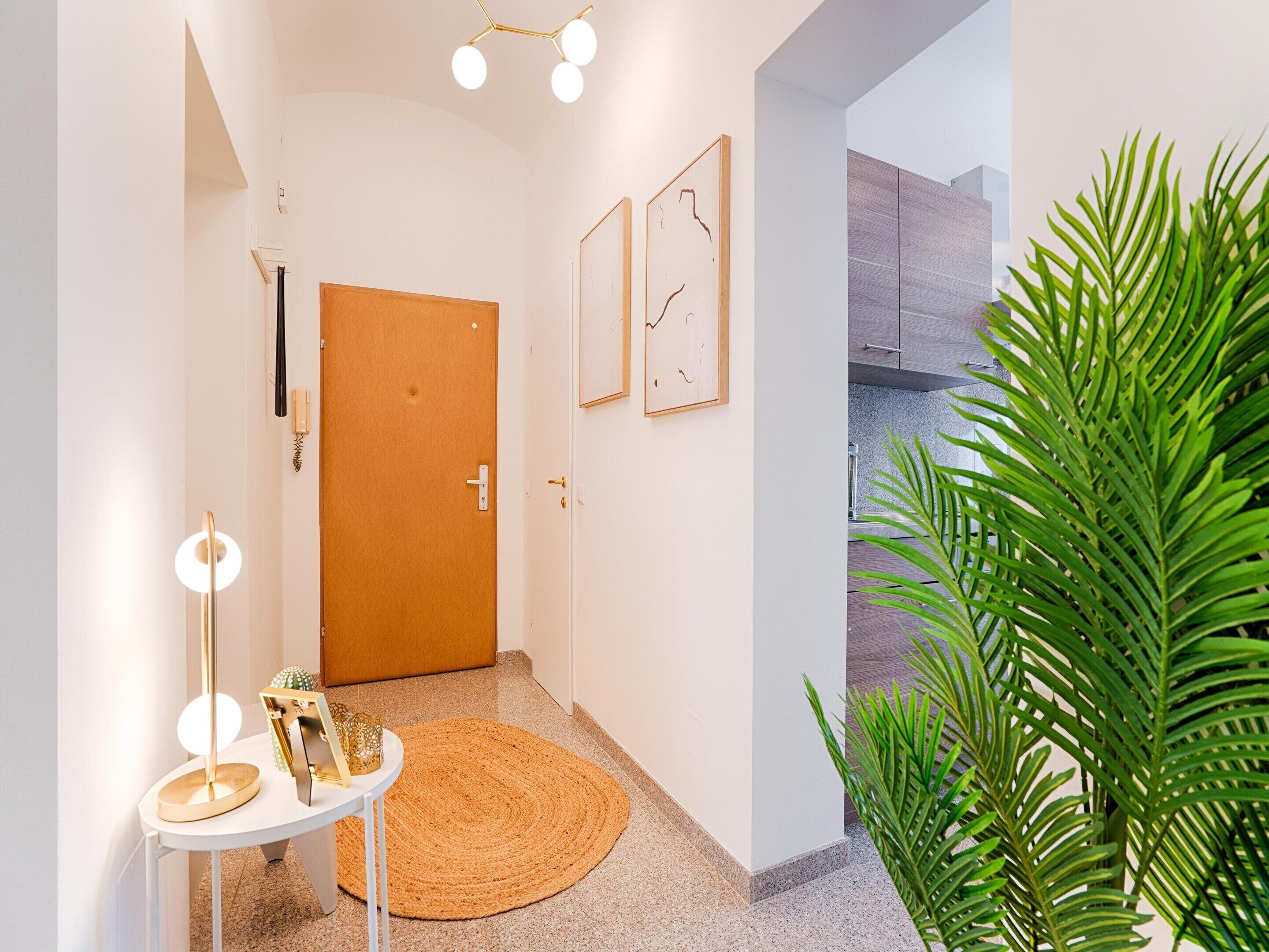 A hallway with a wooden door, a round woven rug, a small table with a lamp, and a potted plant.