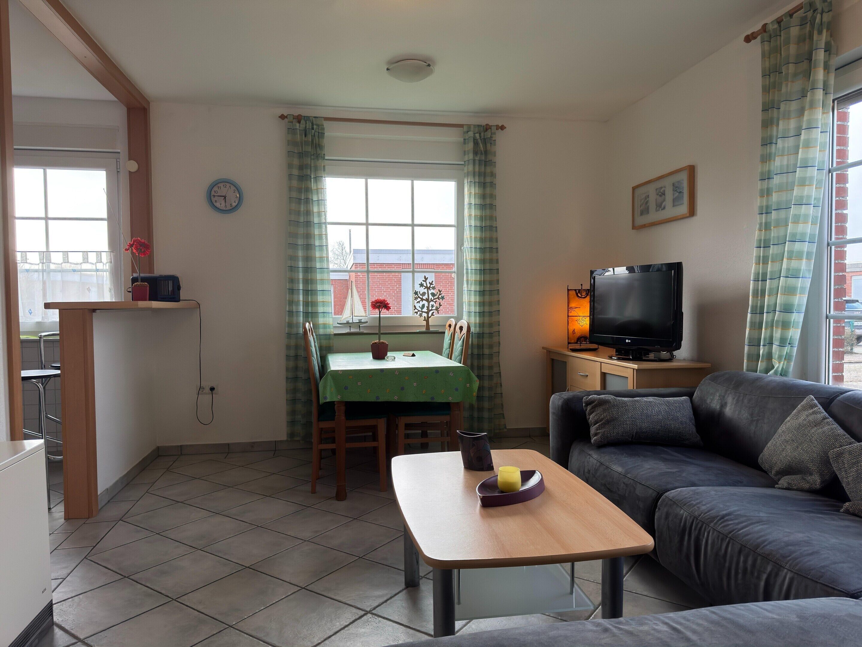 Una sala de estar con sofá, una mesita, televisor, mesa de comedor con sillas y una ventana con cortinas.
