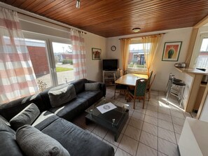 A living room with a sofa, a coffee table, a TV, a dining table with chairs, and a kitchen area with a bar stool. - Bungalow in Strandnaehe, Dornumersiel (Dornum)