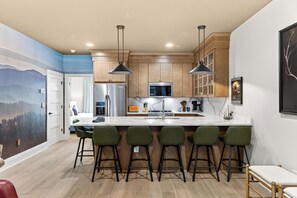 A modern kitchen with a large island, bar stools, and wooden cabinets. - Rooftop Lounge + Hot Tub + Karaoke + Firepit | Katie Hill Collection (Nashville)