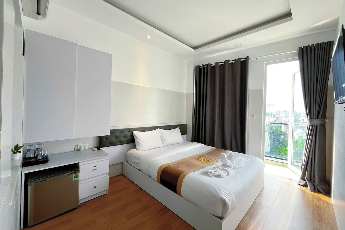 A modern hotel room with a bed, a small desk, a built-in mini-fridge, and a large window with curtains.