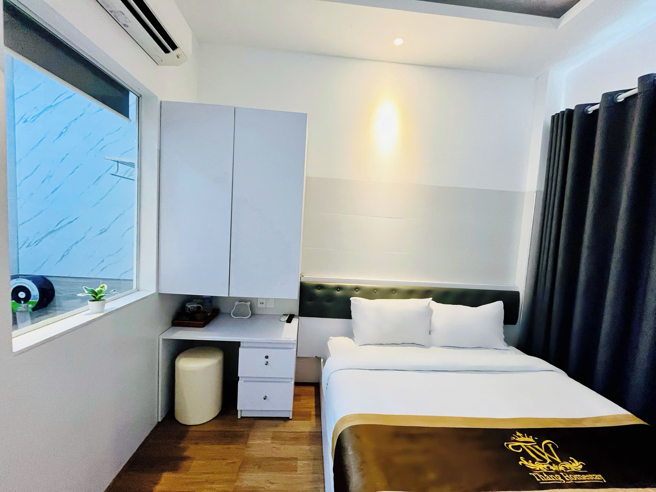 A hotel room with a bed, a bedside table, a small potted plant, a window with a view, and a curtain.