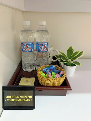A tray with two bottles of water, a mug, a basket of snacks, a potted plant, and a complimentary card. - THANG HOMESTAY (An Hoi Dong)