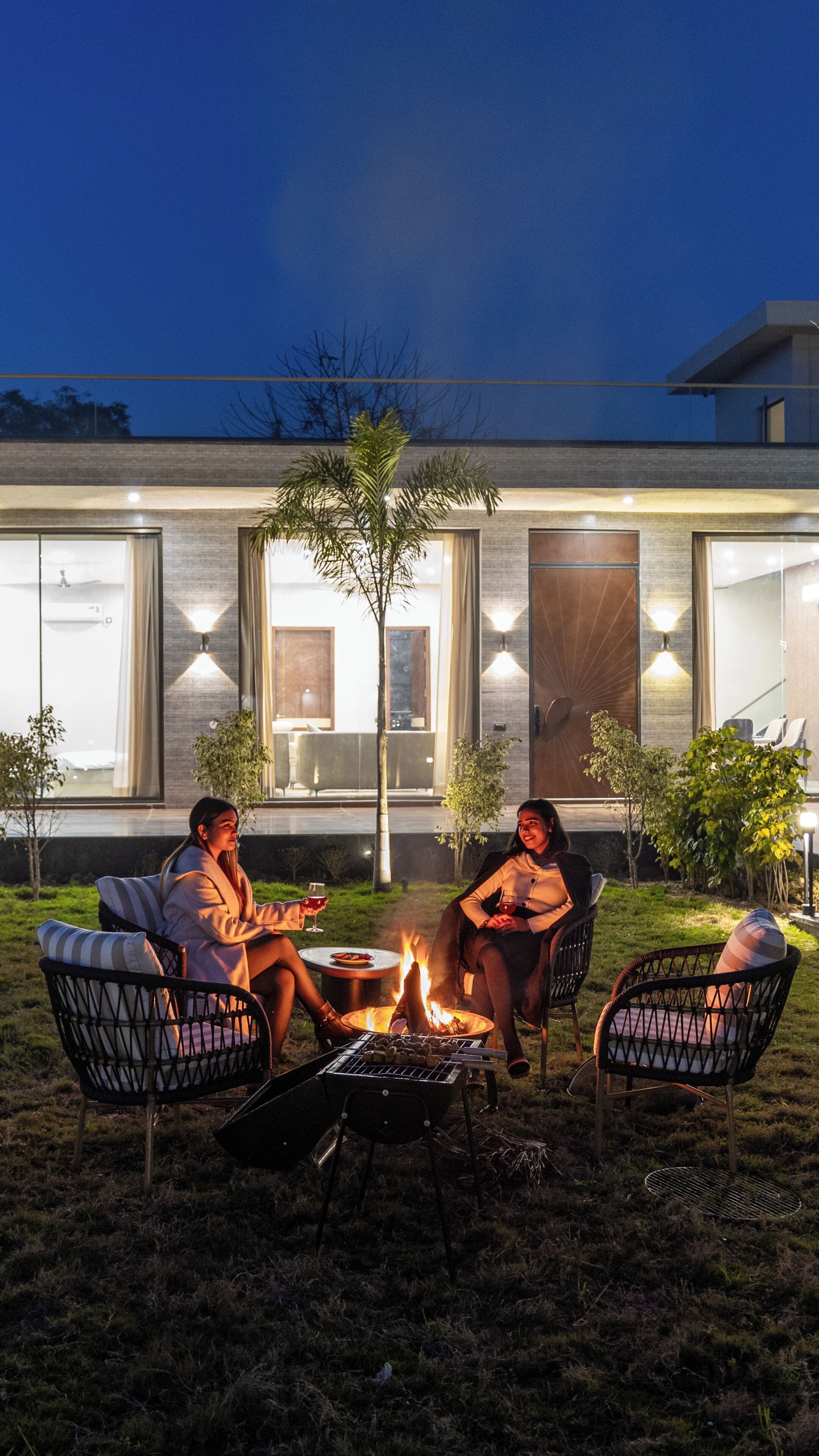 Un espace extérieur aménagé avec des sièges, un foyer et des tables, situé devant un bâtiment moderne doté de grandes fenêtres et d’une entrée bien éclairée.