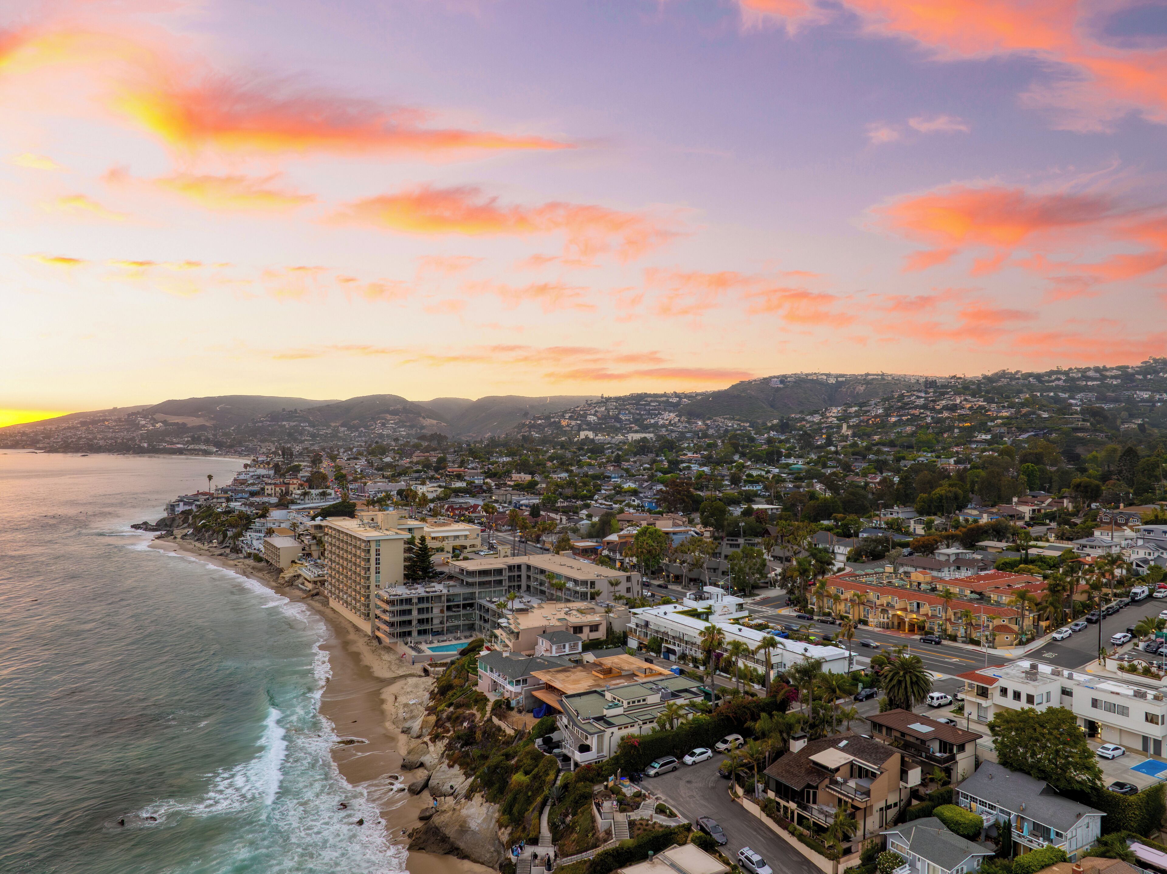 A coastal cityscape with a beach, residential buildings, and a hilly backdrop during sunset.