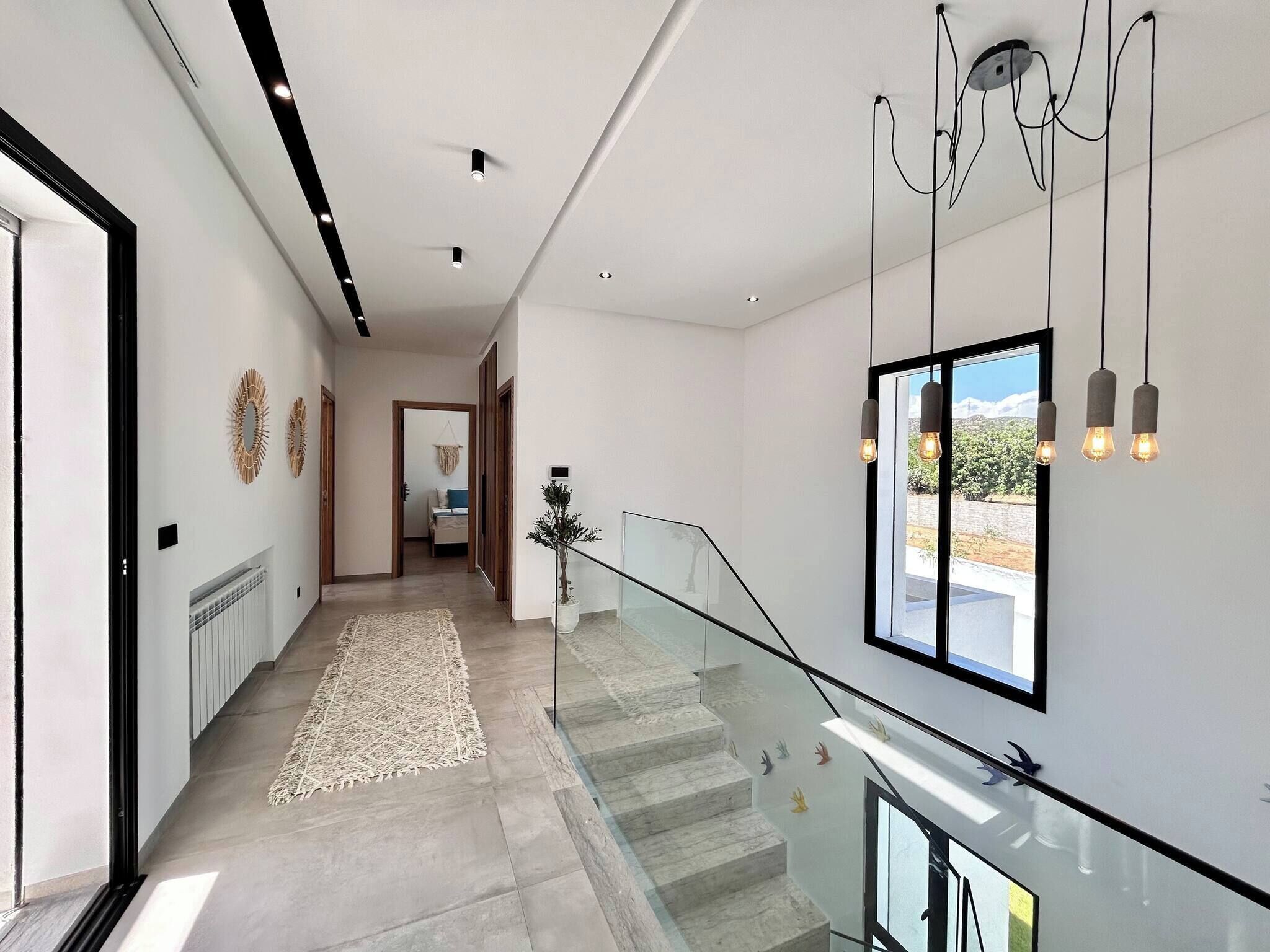 Un intérieur moderne avec un escalier en verre, un couloir menant à une chambre, et une vue dégagée sur l’extérieur grâce à une grande fenêtre.