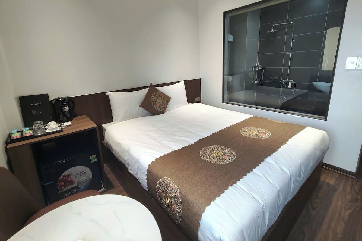 A hotel room with a large bed, a bedside table with a coffee maker, a tray with breakfast items, and a large window with a bathroom view.