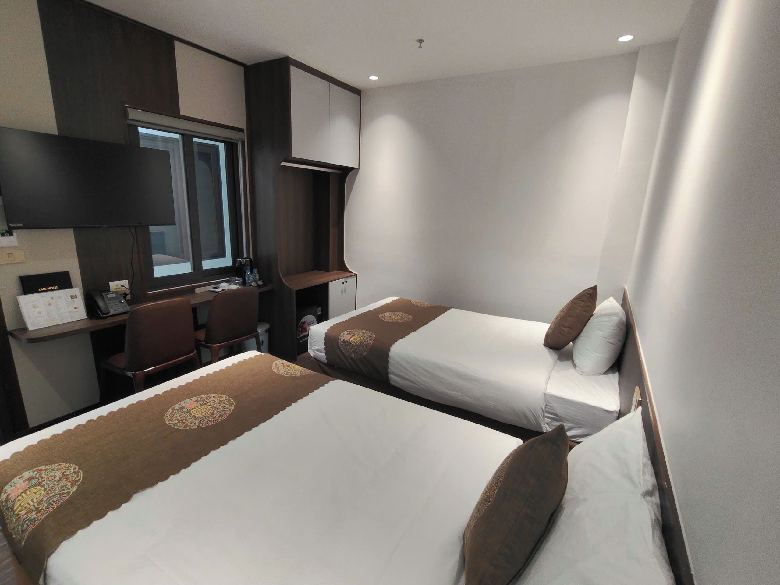 A hotel room with two beds, a desk, a chair, and a window.