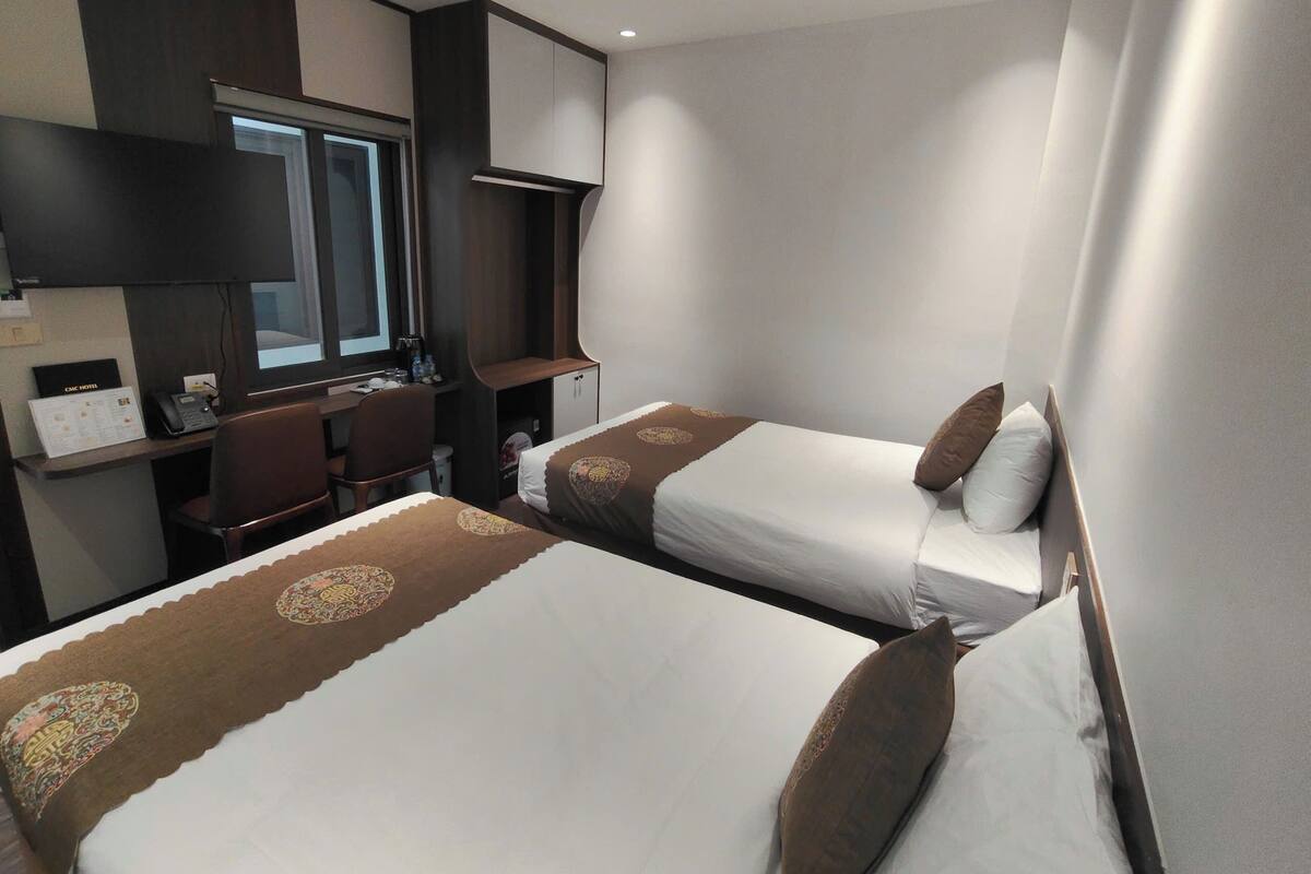 A hotel room with two beds, a desk, a chair, and a window.