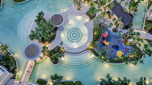 Vue aérienne d’un complexe hôtelier doté d’une piscine centrale, de plusieurs éléments aquatiques et d’espaces paysagers environnants.