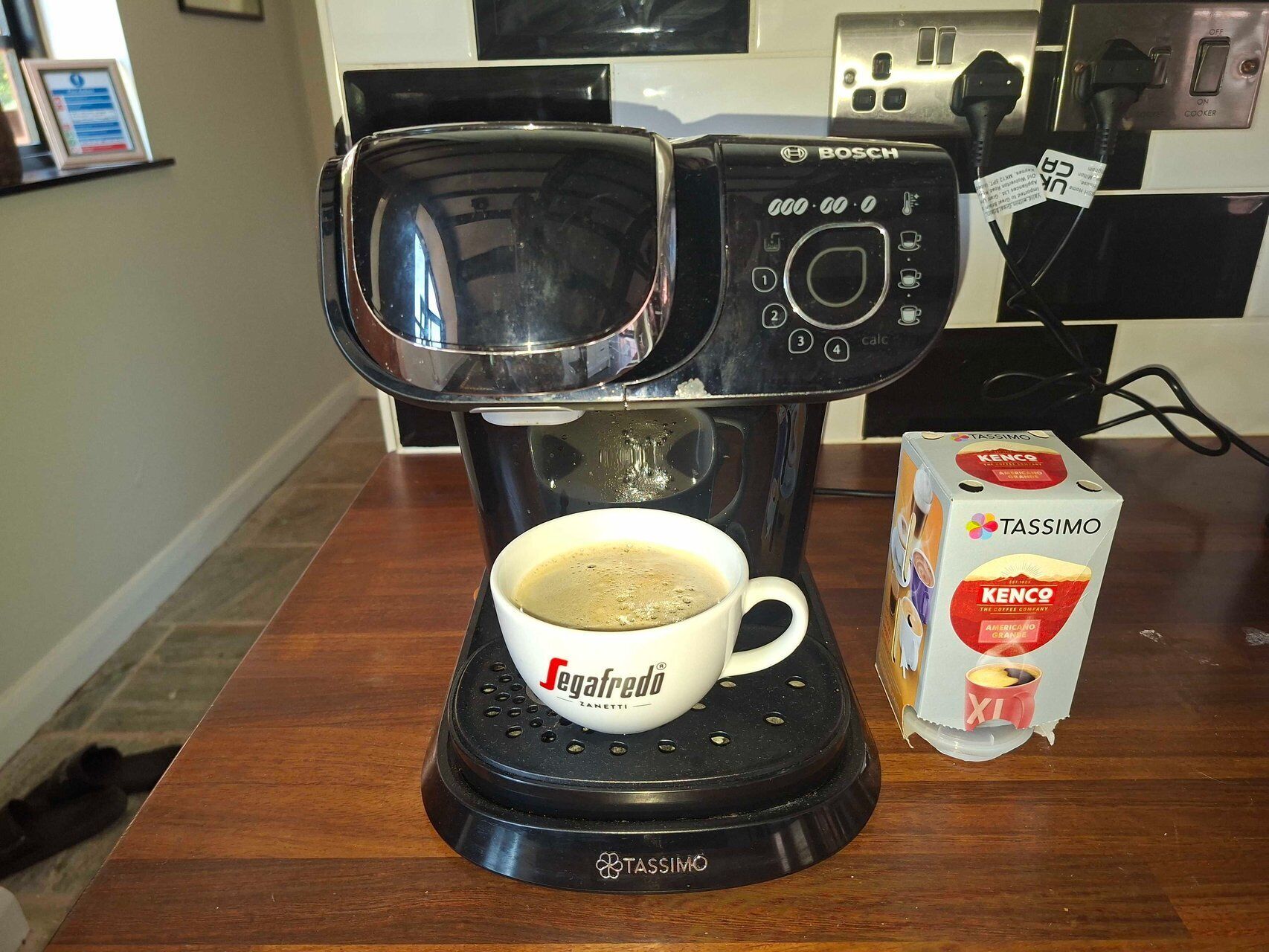 A coffee maker with a cup of coffee on a table, a box of Tassimo pods, and a kitchen appliance in the background.