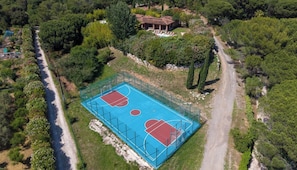 Eine Vogelperspektive zeigt ein Anwesen mit Basketballfeld, Schwimmbad und einem von Bäumen umgebenen Haus.
