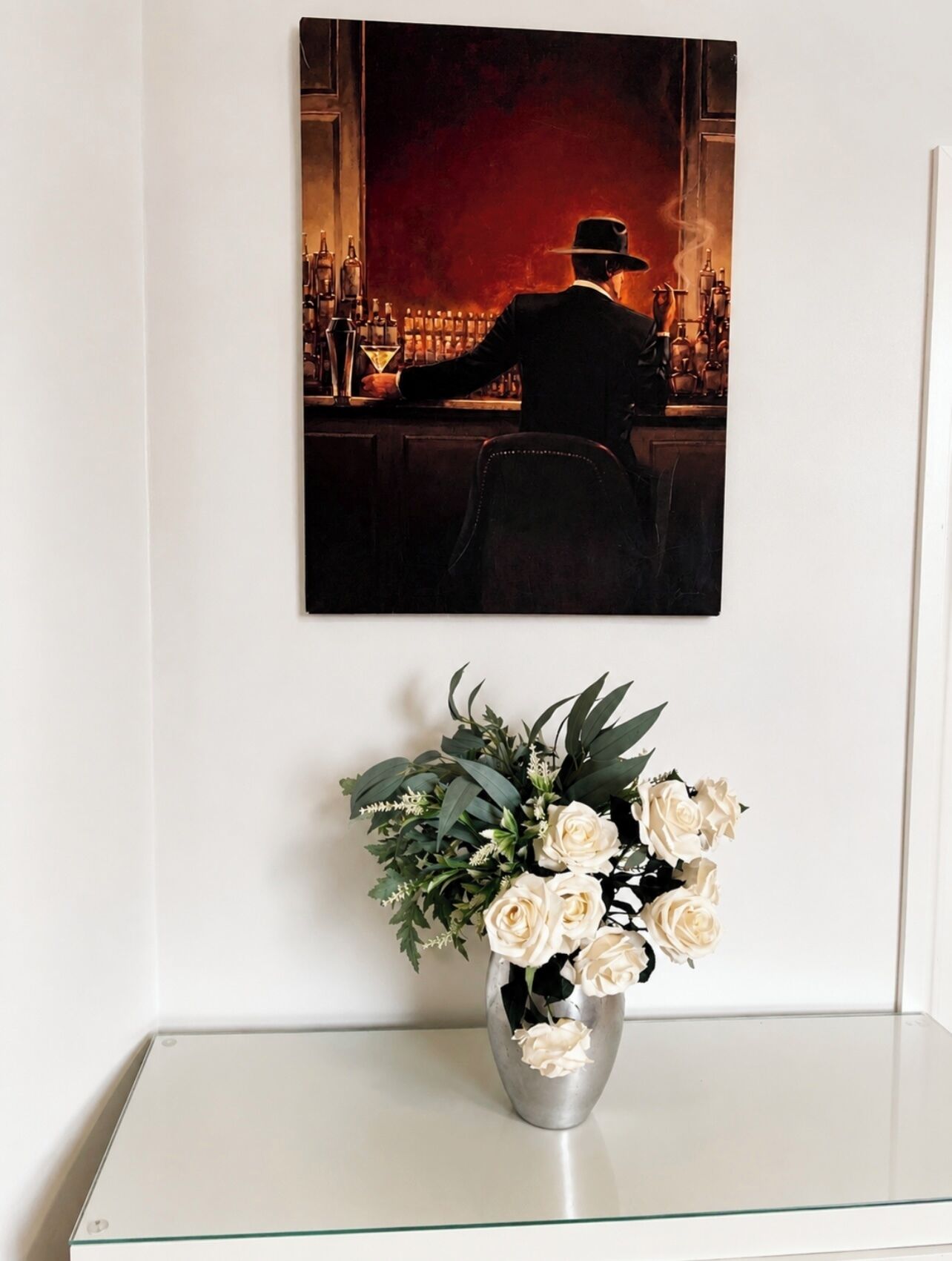 A framed painting of a bar scene with a person in a suit and hat, a bar counter with bottles, and a vase with white flowers on a white shelf.