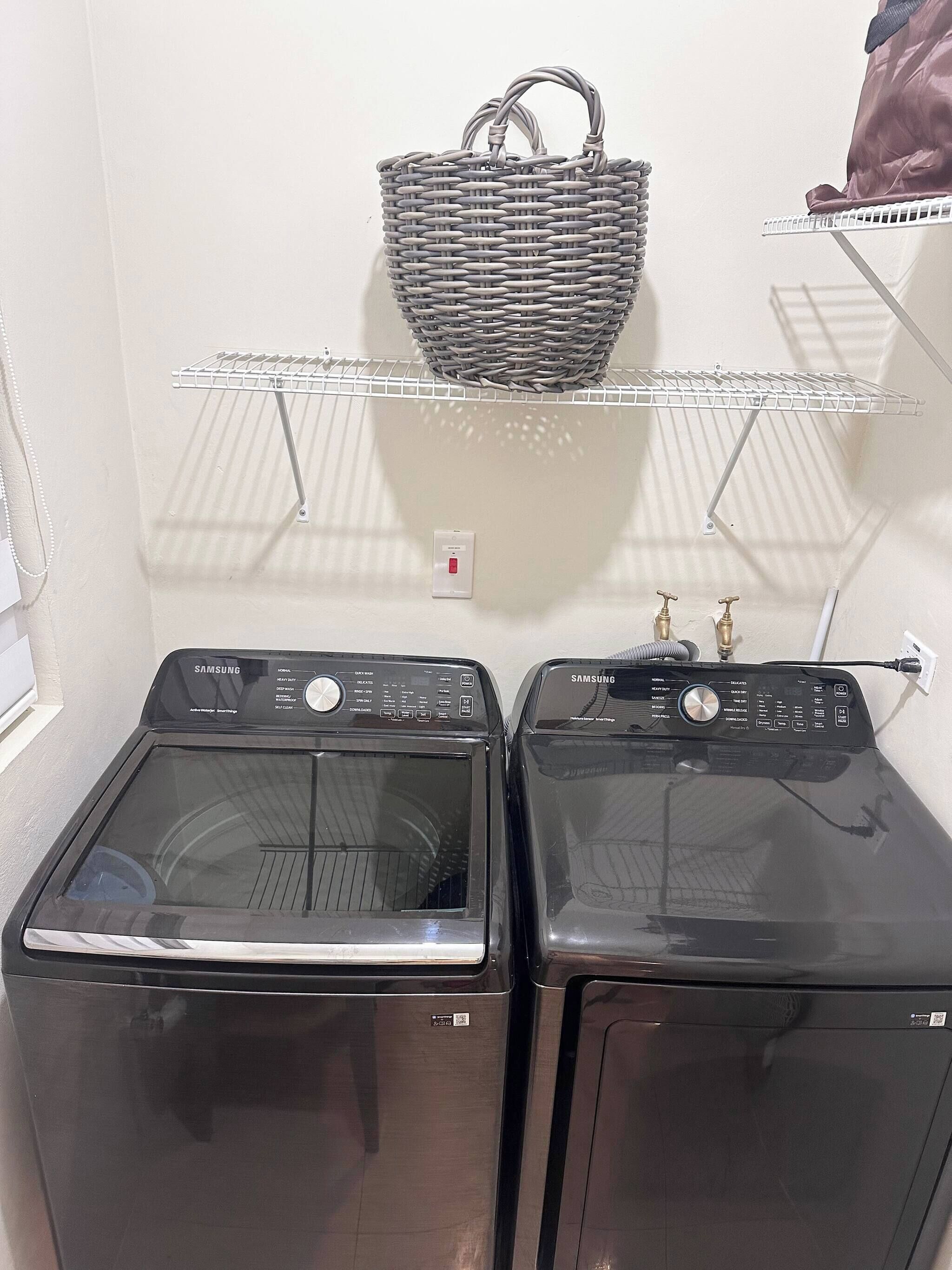Deux machines à laver noires dotées de panneaux de commande, un mur blanc, un panier suspendu et une étagère blanche.