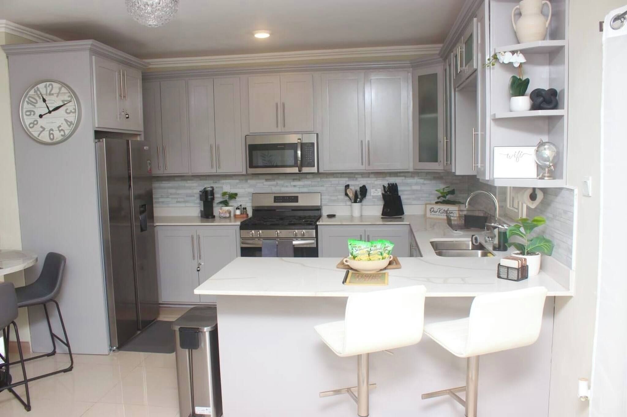Una cocina moderna con armarios blancos, electrodomésticos de acero inoxidable, una isla blanca con taburetes de bar y un reloj en la pared.