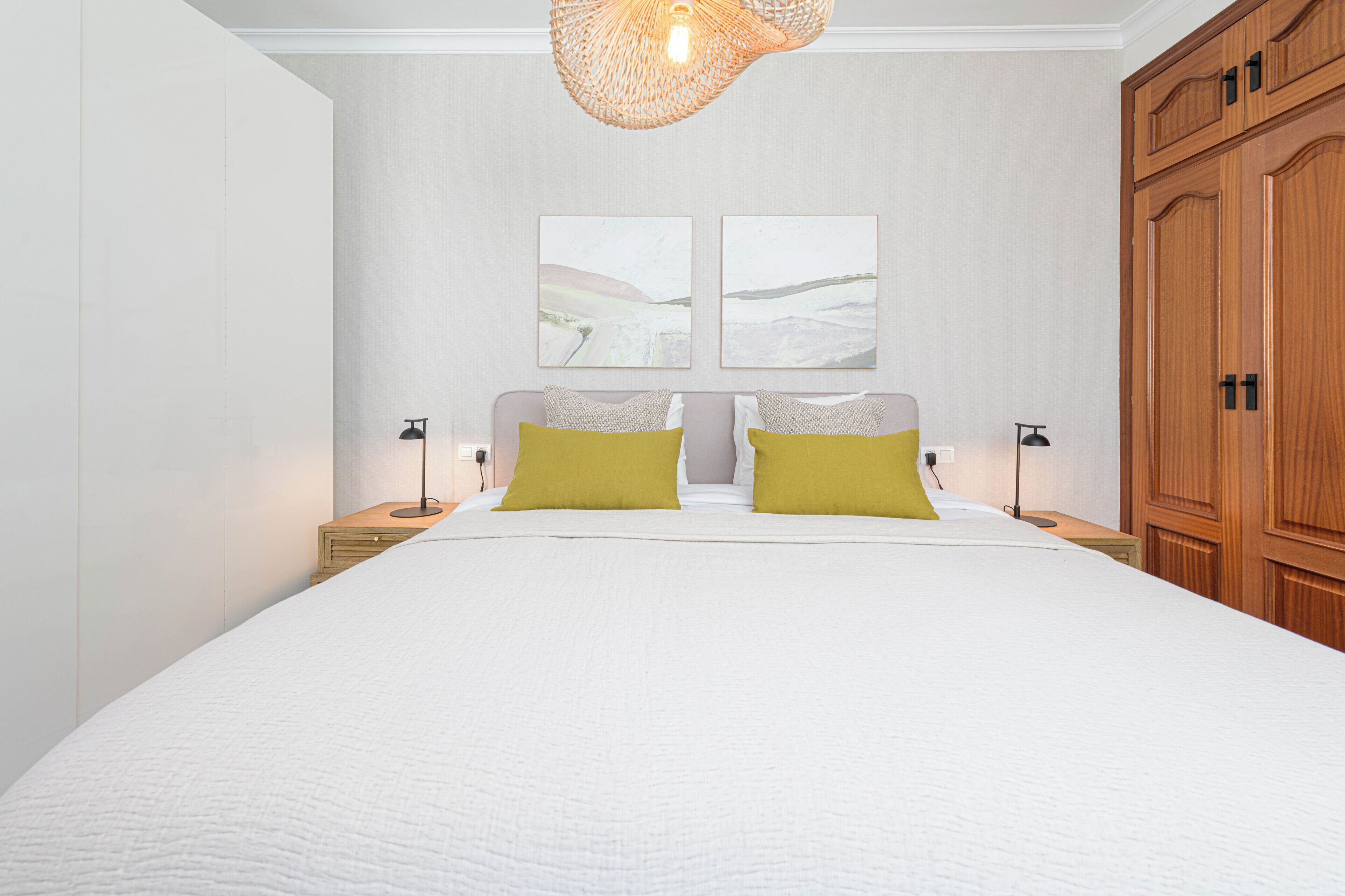 A bedroom with a large bed, two bedside tables, a wooden wardrobe, and two wall-mounted artworks.