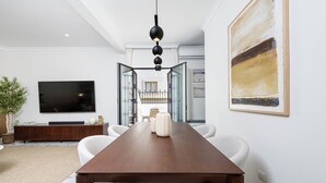 Una sala da pranzo moderna con un lungo tavolo in legno, sedie bianche, un televisore a schermo piatto e un grande dipinto astratto appeso alla parete.