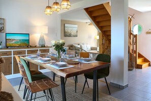 A dining area with a wooden table set for four, surrounded by wicker chairs, a vase with flowers, and a television displaying a scenic view. - Cancal'ou (Cancale)