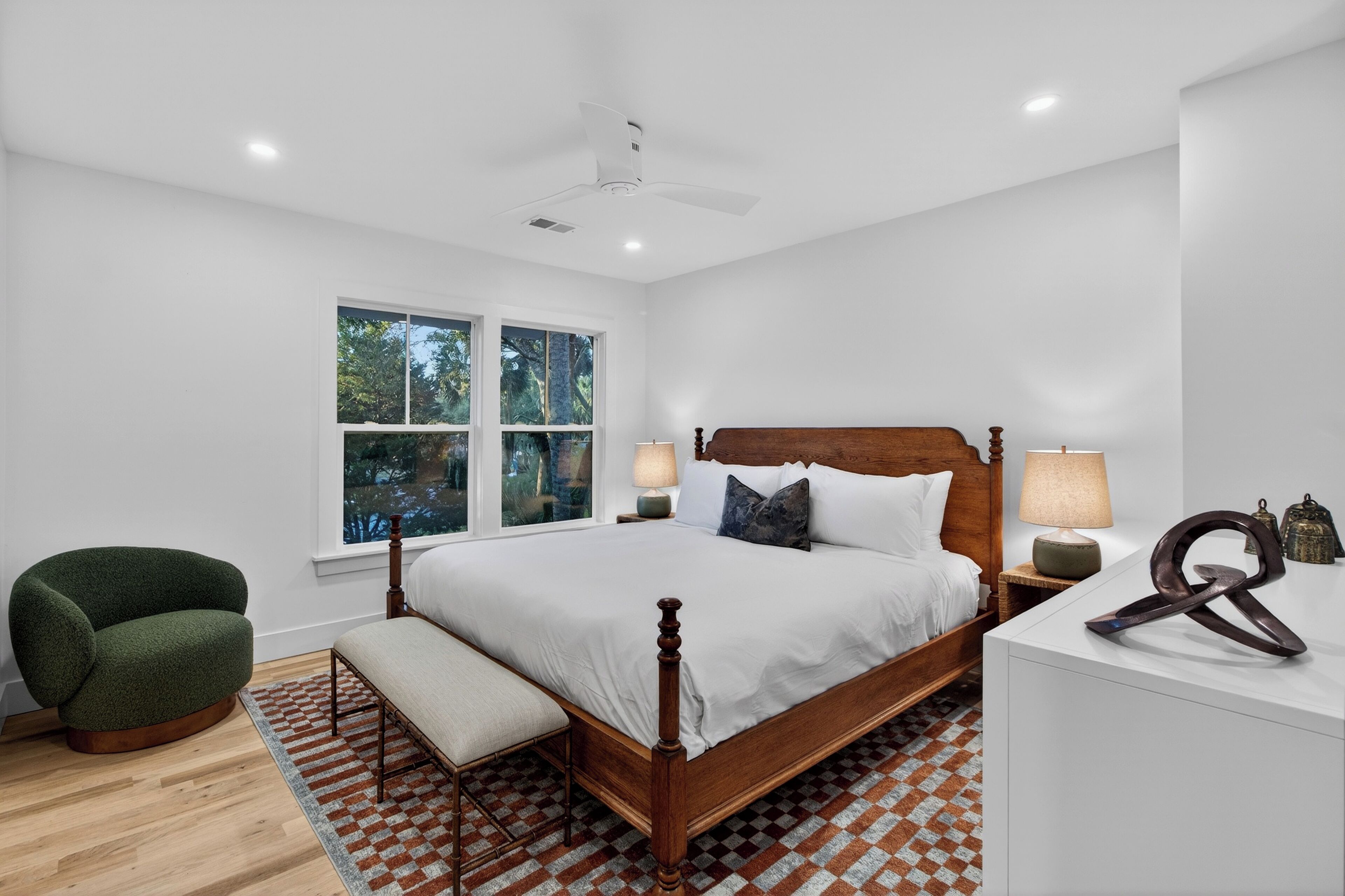 A bedroom with a wooden bed, a green armchair, a bench, a patterned rug, and a window with a view of trees.