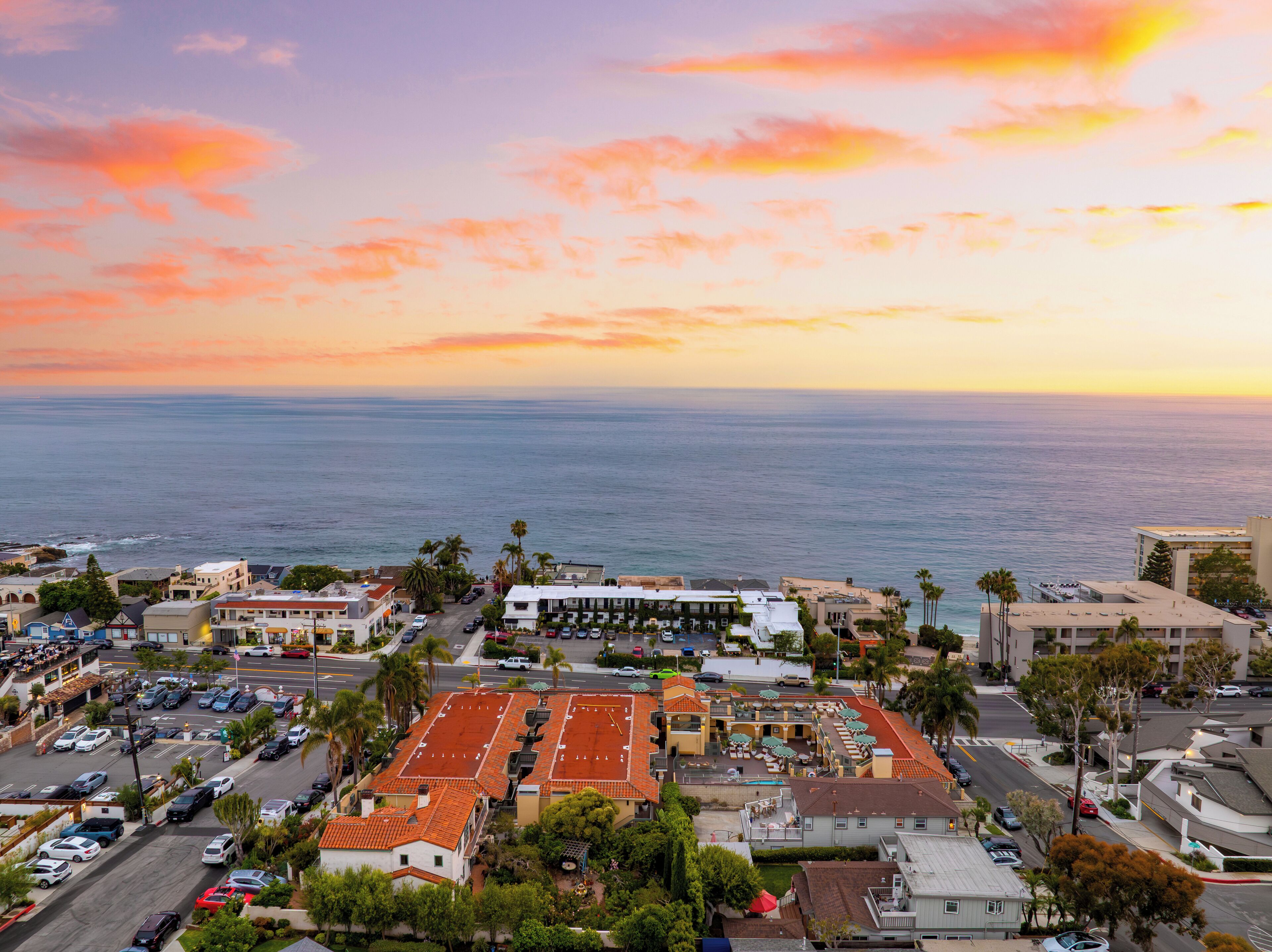 A coastal town with a prominent building featuring a red-tiled roof, surrounded by parked cars and palm trees.