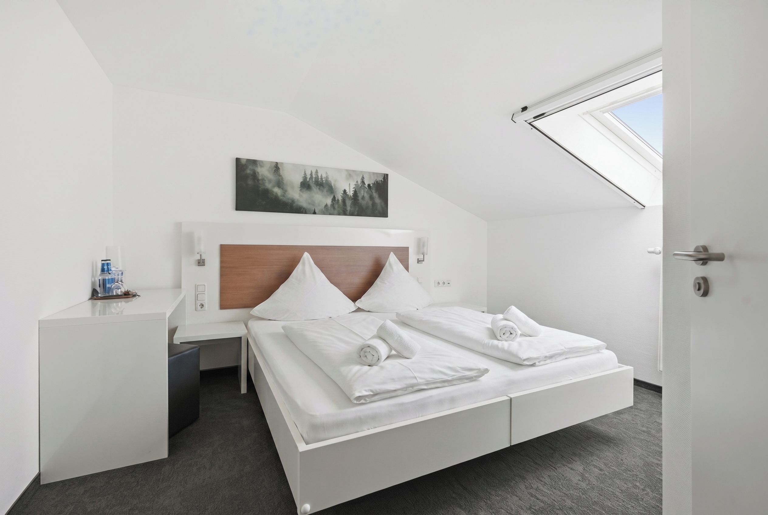 A modern hotel room with a bed, a desk, a painting, and a skylight.