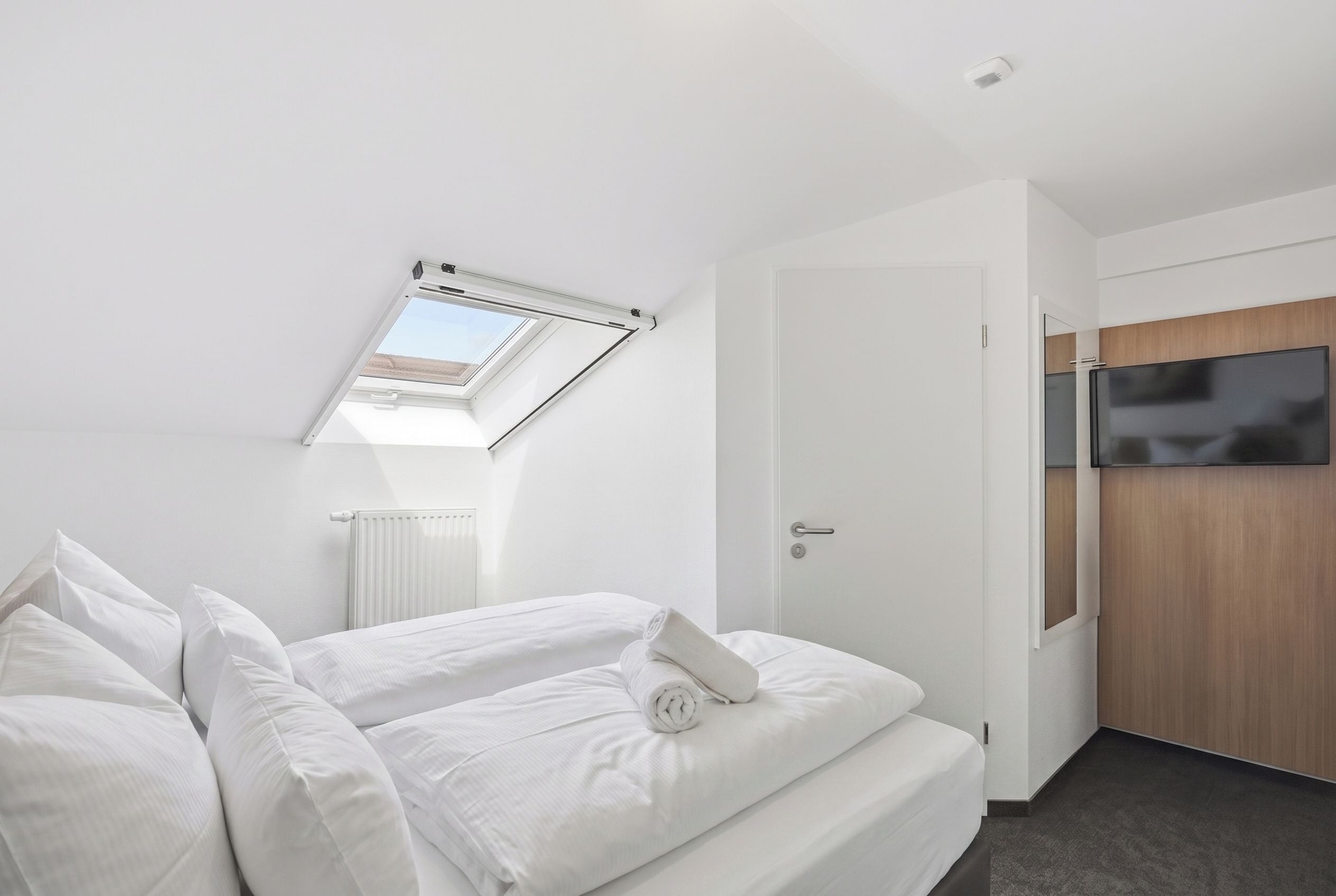 Een moderne slaapkamer met dakraam, een bed met witte lakens, een radiator en een televisie.