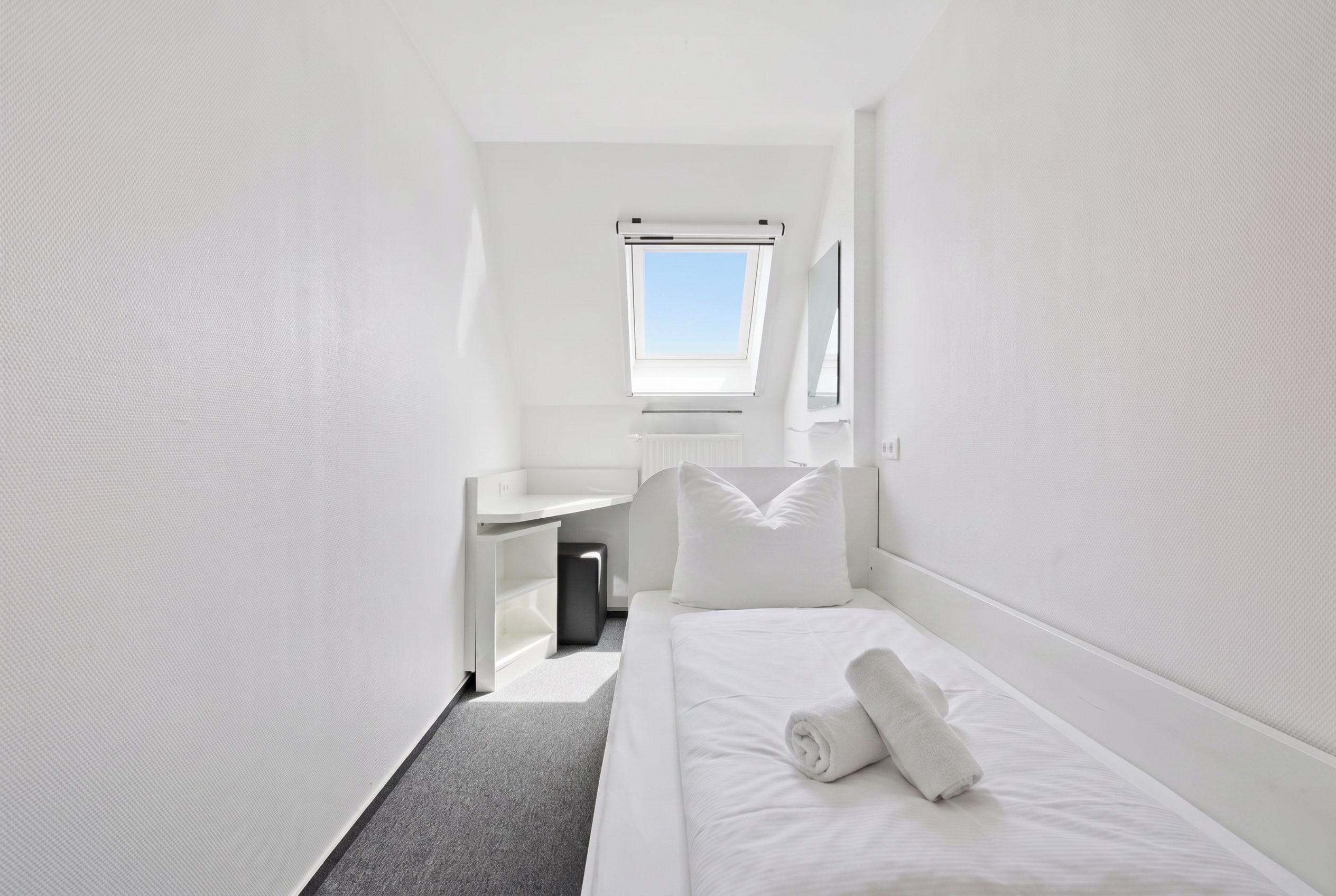 A small, white, single-room hotel room with a bed, a desk, and a window.