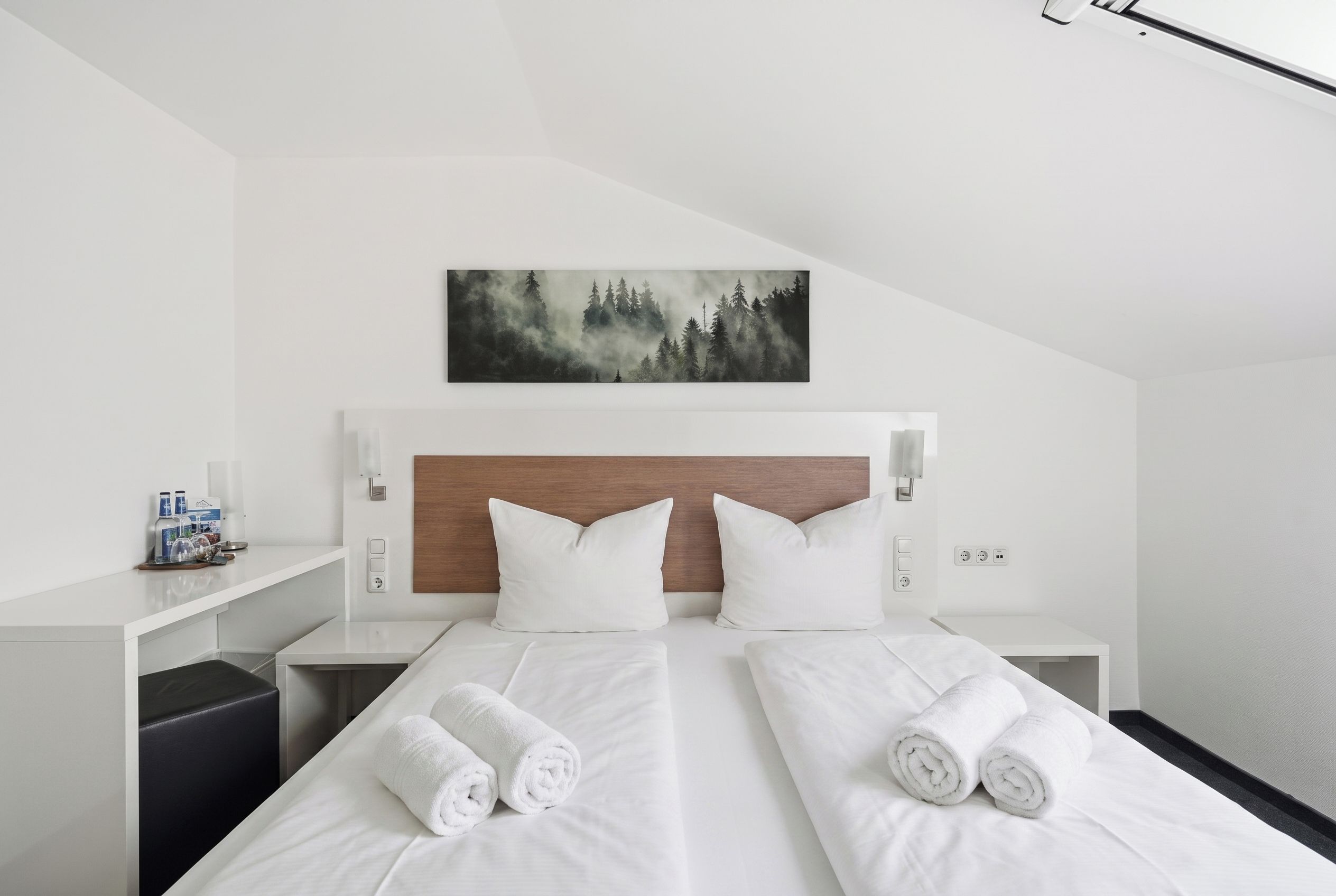 Habitación de hotel con una cama grande, dos almohadas, cabecero, mesita de noche con una botella y vasos, y un cuadro de un bosque.