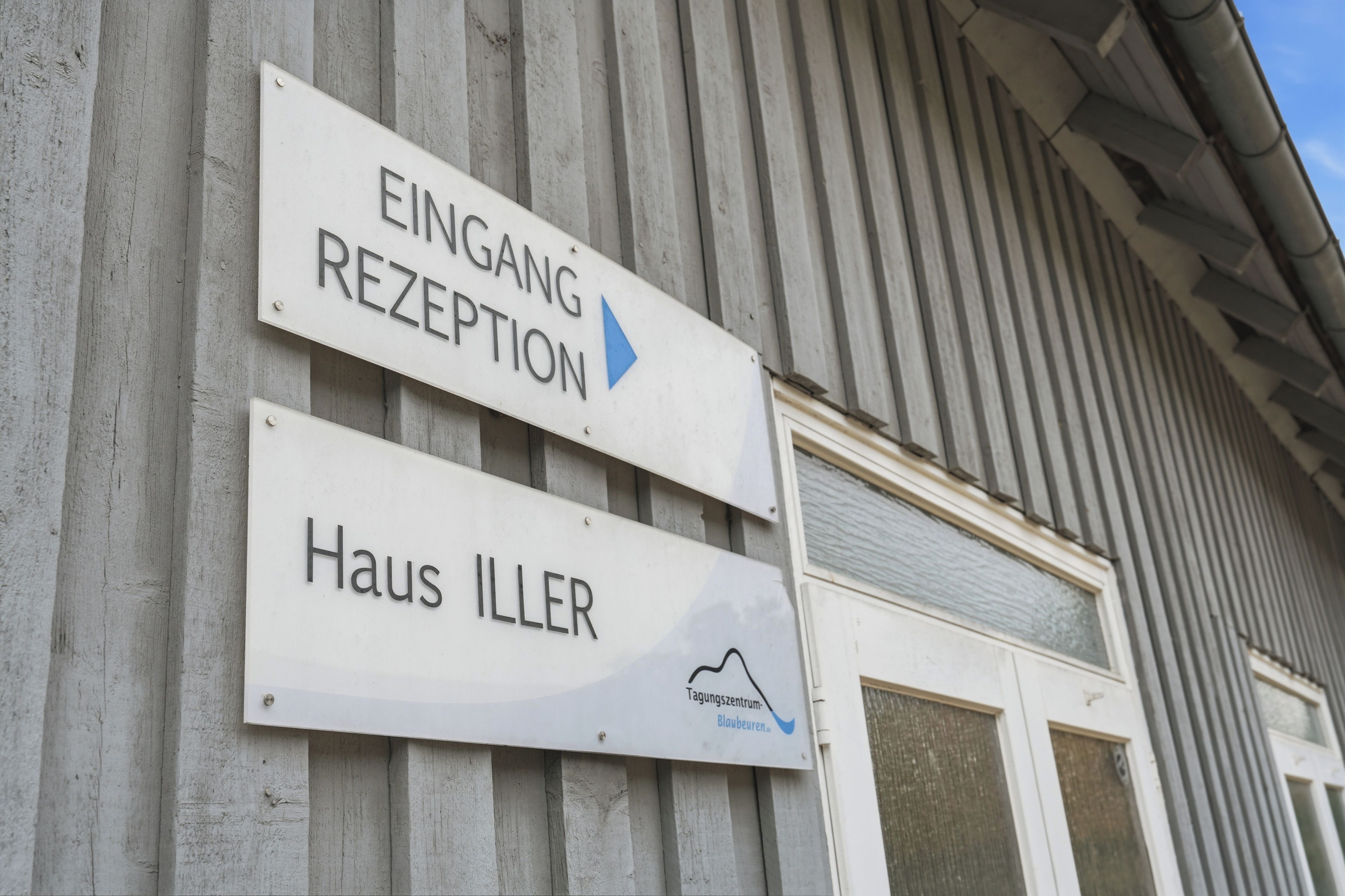 A building with two signs: one indicating 'EINGANG REZEPTION' with an arrow pointing to the entrance, and another sign for 'Haus ILLER' with a logo below.