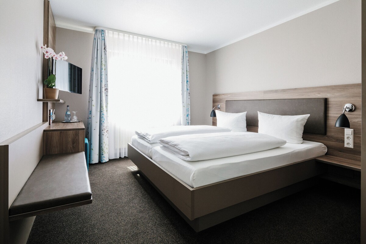 Une chambre d’hôtel avec un grand lit, une table de chevet, un miroir, une fenêtre avec des rideaux et une petite plante.