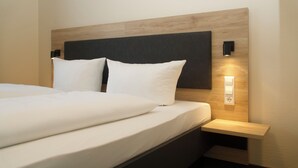 A neatly made bed with white linens and pillows, a wooden headboard, a bedside table with a lamp, and a wall-mounted light fixture. - Hotel & Tagungszentrum Blaubeuren (Blaubeuren)