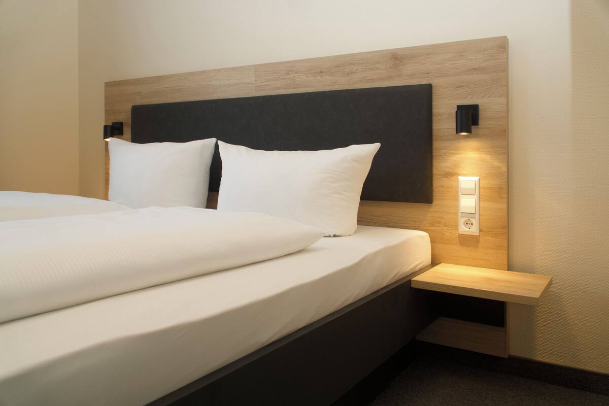 Ein ordentlich bezogenes Bett mit weißen Leinen und Kissen, ein hölzerner Kopfteil, ein Nachttisch mit Lampe und eine Wandleuchte.