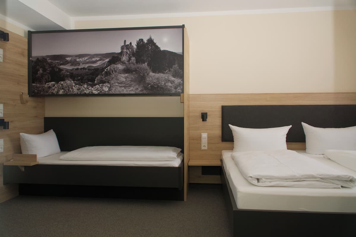 Ein Hotelzimmer mit einem großen Bett, einem kleineren Bett, einem Nachttisch, einem an der Wand befestigten Fernseher und einem Landschaftsbild.