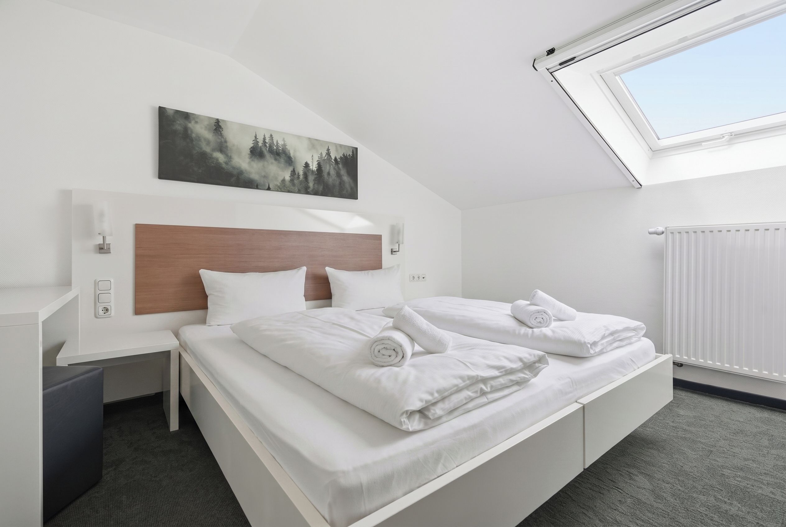 A hotel room with a bed, a nightstand, a wall-mounted lamp, a painting, and a skylight.