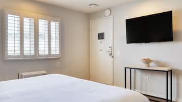 A bedroom with a bed, a TV mounted on the wall, a window with blinds, and a radiator.