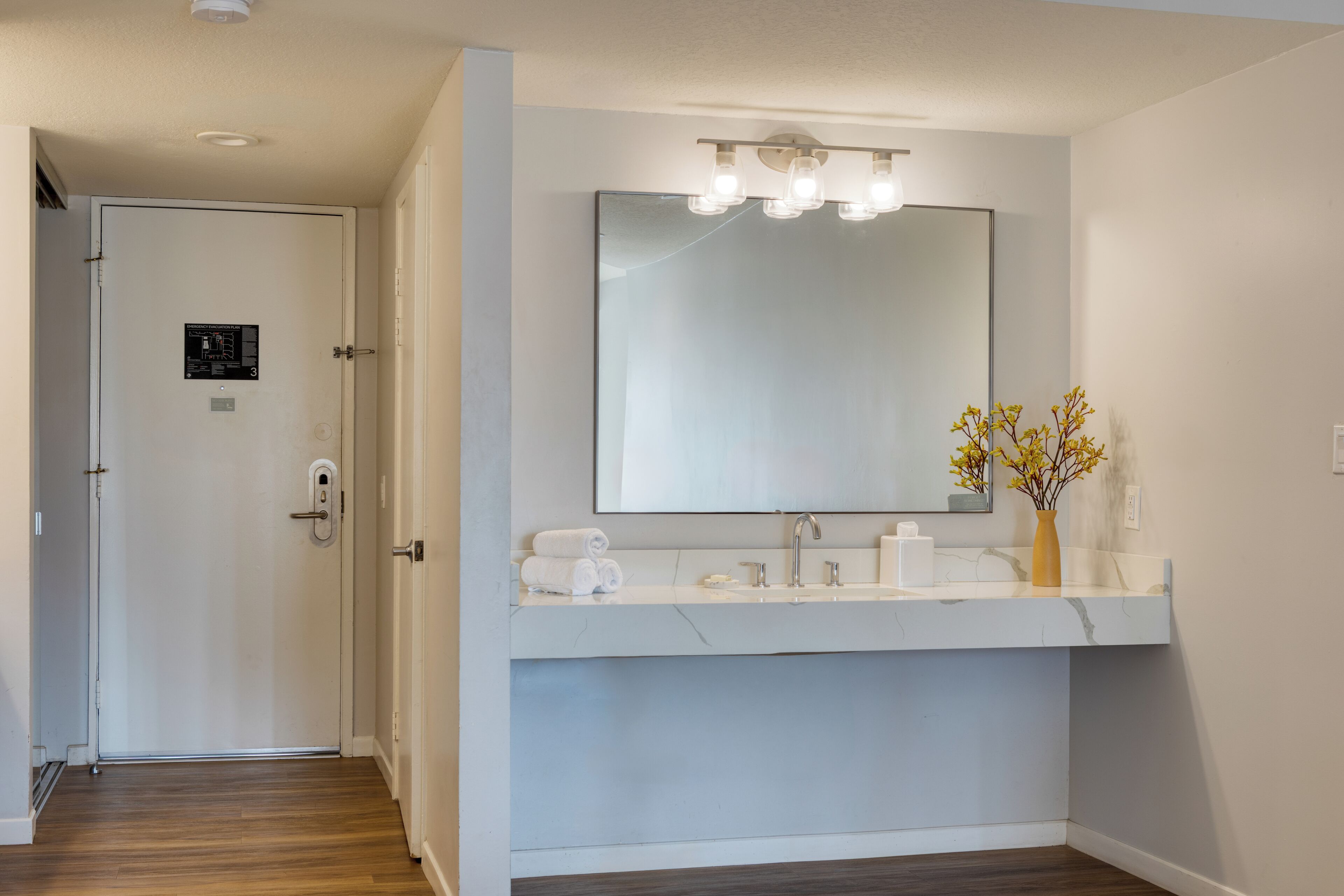 Une salle de bain avec un lavabo blanc, un miroir et un vase contenant des fleurs jaunes.