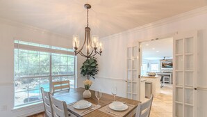 A dining room with a wooden table, white chairs, a chandelier, and a view of a pool through large windows. - Resort-Style Pool + Spa + BBQ. Spacious 4 bdr Home @The Woodlands (Spring)