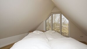 Quarto com teto inclinado, uma janela grande e cama com roupa de cama branca.