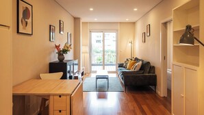 Uma sala de estar moderna com sofá, mesa de centro e televisão, uma área de jantar com mesa e cadeiras, e uma grande porta de vidro deslizante para o exterior.