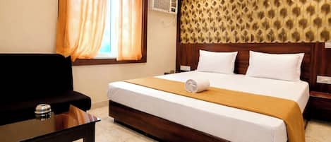 A hotel room with a bed, a desk, a chair, a window with curtains, an air conditioning unit, and a patterned headboard.