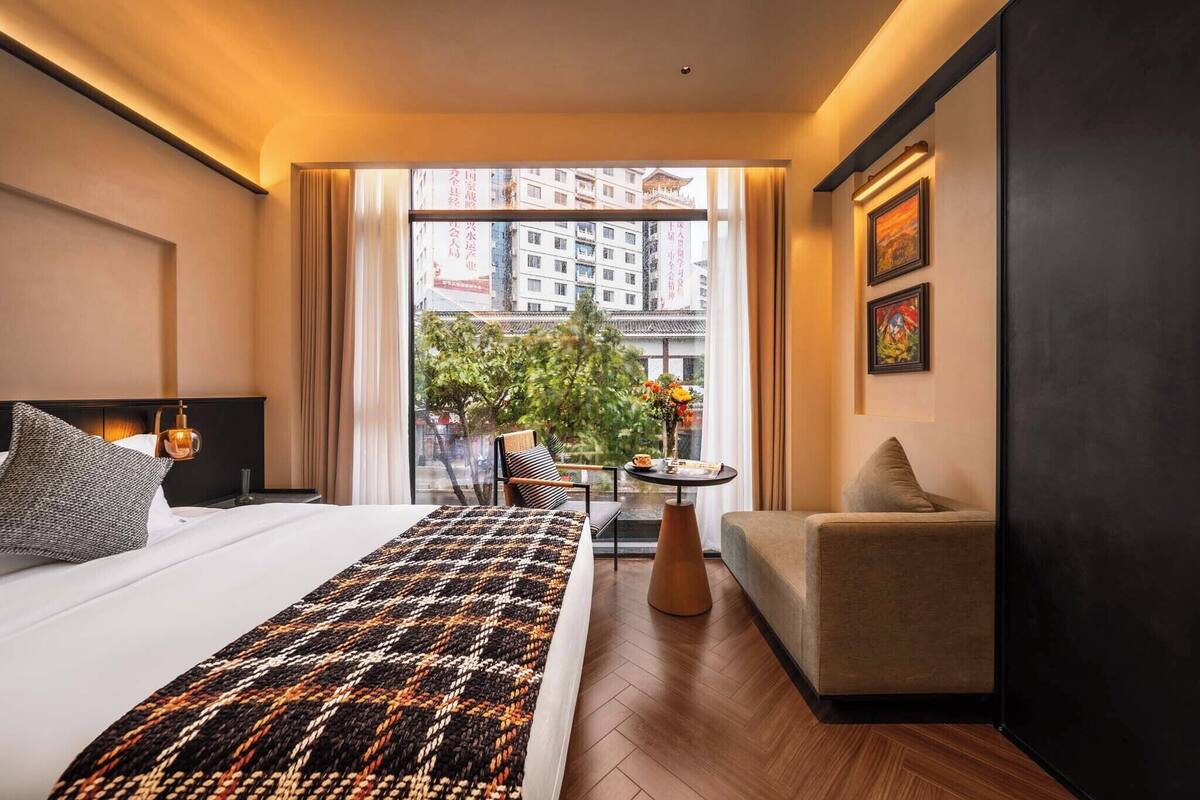 Une chambre d’hôtel moderne, dotée d’un grand lit, d’un canapé, d’une petite table et offrant une vue sur la ville grâce à de grandes fenêtres.