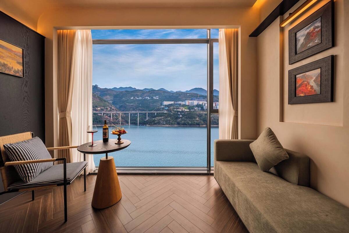 Une chambre d’hôtel moderne avec une grande fenêtre, un canapé, un fauteuil, une petite table avec une bouteille et des fruits, et deux tableaux encadrés au mur.